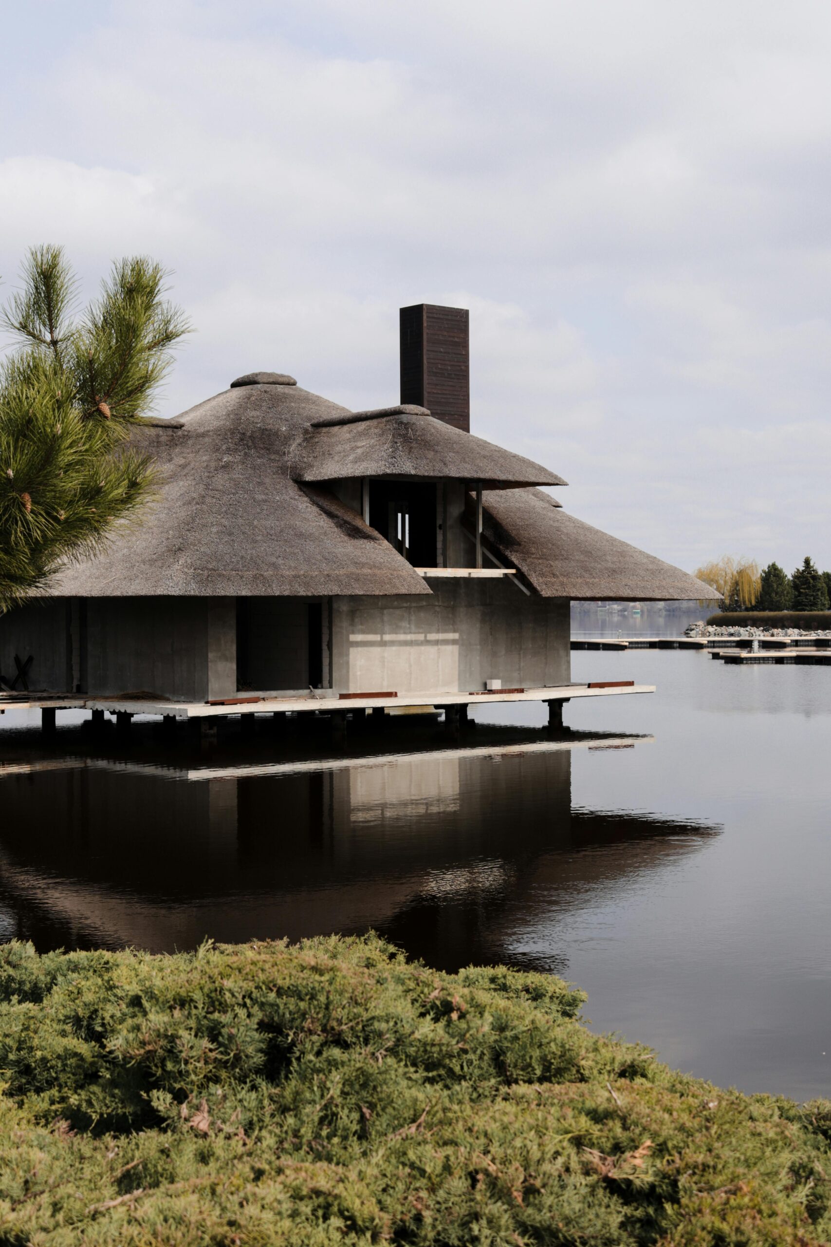 A peaceful lake house with a unique thatched roof reflected on still water, surrounded by nature.
